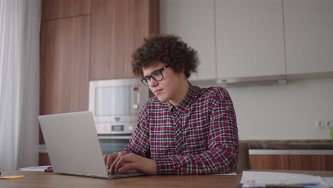 A-Curly-man-with-a-serious-look-works-at-a-laptop-sitting-in-a-modern-kitchen.-Young-man-freelancer-student-using-laptop-studying-online-working-from-home-in-internet